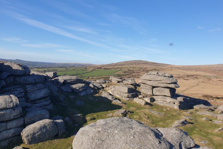A view of Dartmoor from one of the Tors. In the foreground are towers of grey rock, with the moor behind and another tor visible in the distance. The sky is a clear deep blue with some streaks of white cloud across it.