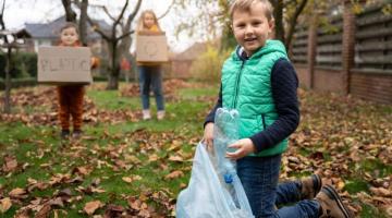 Three children collecting litter in a park. Two of them hold cardboard boxes and the third, closest to the camera has a plastic bag.