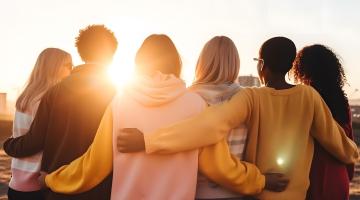 A photo of six young people, with their backs to us, looking into a bright sunrise. The six people have their arms locked around each other, in a gentle uniting embrace. They are all wearing different coloured jumpers. 
